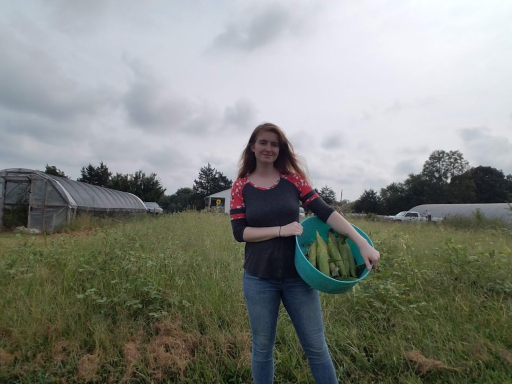 This image displays me a few years ago on my family's farm holding a bucket of freshly picked corn in front of a grassy field and a hoop house.